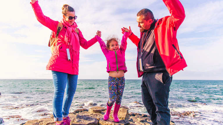 Family of three (two parents, one child) standing on the Grand Causeway with their arms in the air, enjoying a GIANT day out at Northern Ireland's iconic World Heritage Site
